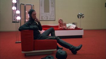 Movie still from “Death Race 2000” (1975), directed by Paul Bartel – A man sitting on top of a red couch next to a woman; Wide shot, Low angle