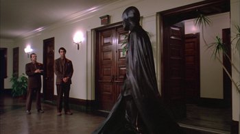 Movie still from “Death Race 2000” (1975), directed by Paul Bartel – A man in a black costume walking in front of a door way; Wide shot, Over the shoulder angle
