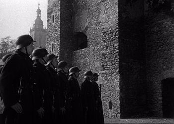 Movie still from “Decision Before Dawn” (1951), directed by Anatole Litvak – A black and white photo of a group of men standing next to each other in front of a stone wall; Wide shot, Low angle