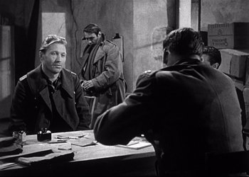 Movie still from “Decision Before Dawn” (1951), directed by Anatole Litvak – Two men are sitting at a table and one of the men is holding a gun; Medium shot, Over the shoulder angle