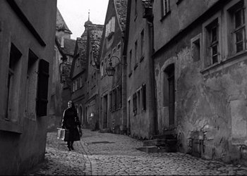 Movie still from “Decision Before Dawn” (1951), directed by Anatole Litvak – An old woman walking down a street with a bag; Wide shot, High angle