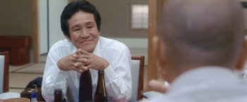 Movie still from “Demonlover” (2002), directed by Olivier Assayas – A man sitting at a table with his hands clasped together; Medium shot, Low angle