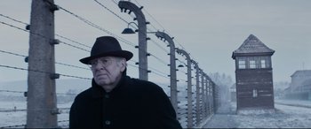 Movie still from “Denial” (2016), directed by Mick Jackson – An old man wearing a black hat standing in front of barbed wire; Medium shot, Over the shoulder angle