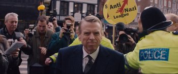 Movie still from “Denial” (2016), directed by Mick Jackson – A man in a suit and tie standing in front of a group of onlookers; Medium shot, Over the shoulder angle