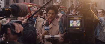 Movie still from “Denial” (2016), directed by Mick Jackson – A woman is holding a microphone while standing in front of a group of onlookers; Medium shot, Over the shoulder angle