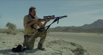 Movie still from “Desierto” (2015), directed by Jonás Cuarón – A man kneeling down holding a rifle in the desert; Medium shot, Low angle