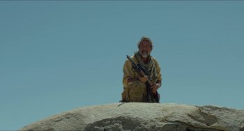 Movie still from “Desierto” (2015), directed by Jonás Cuarón – A man sitting on top of a large rock holding a rifle; Medium shot, Low angle