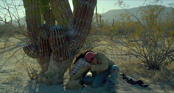 Movie still from “Desierto” (2015), directed by Jonás Cuarón – A man sitting on the ground next to a palm tree; Wide shot, Overhead angle