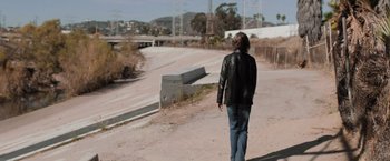 Movie still from “Destroyer” (2018), directed by Karyn Kusama – A man walking down a dirt road near a bridge; Wide shot, Over the shoulder angle