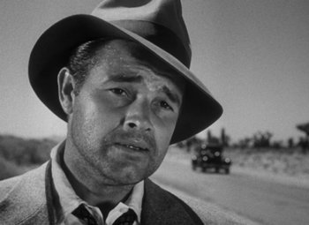 Movie still from “Detour” (1945), directed by Edgar G. Ulmer – A man wearing a hat standing on the side of a road; Close Up shot, Over the shoulder angle
