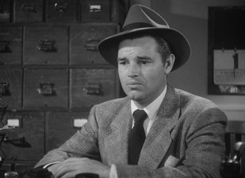 Movie still from “Detour” (1945), directed by Edgar G. Ulmer – A man wearing a suit and a hat sitting in front of a stack of filing cabinets; Close Up shot, Low angle