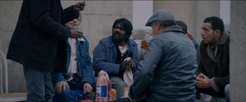 Movie still from “Dheepan” (2015), directed by Jacques Audiard – A group of people sitting around a building eating; Medium shot, Over the shoulder angle