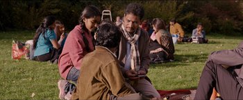 Movie still from “Dheepan” (2015), directed by Jacques Audiard – A group of people sitting in the grass looking at a phone; Medium shot, Over the shoulder angle