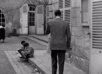Movie still from “Diabolique” (1955), directed by Henri-Georges Clouzot – A man standing on the side of the street next to another man sitting on the sidewalk; Wide shot, Over the shoulder angle