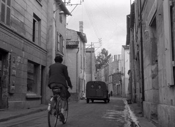 Movie still from “Diabolique” (1955), directed by Henri-Georges Clouzot – A man riding a bike down a street past buildings; Extreme Wide shot, High angle