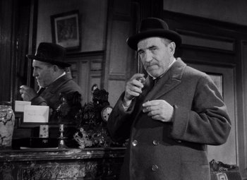 Movie still from “Diabolique” (1955), directed by Henri-Georges Clouzot – A man in a suit and a hat smoking a cigarette; Medium shot, Low angle