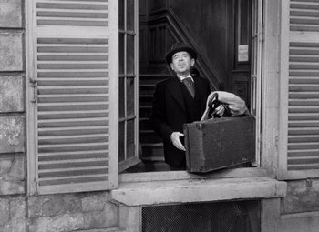 Movie still from “Diabolique” (1955), directed by Henri-Georges Clouzot – A man in a suit and hat holding an old suitcase; Medium shot, Low angle