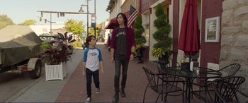 Movie still from “Diary of a Wimpy Kid: The Long Haul” (2017), directed by David Bowers – Two young men walking down a sidewalk near a table; Wide shot, Over the shoulder angle
