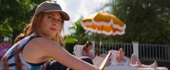 Movie still from “Do Revenge” (2022), directed by Jennifer Kaytin Robinson – A woman sitting in a chair reading a book with an umbrella in the background; Medium shot, Over the shoulder angle