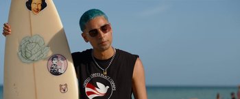Movie still from “Do Revenge” (2022), directed by Jennifer Kaytin Robinson – A man with blue hair holding a surfboard in front of the ocean; Medium shot, Over the shoulder angle