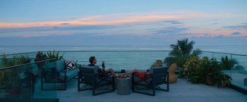 Movie still from “Do Revenge” (2022), directed by Jennifer Kaytin Robinson – Two people sitting in chairs on a deck overlooking the ocean; Wide shot, High angle