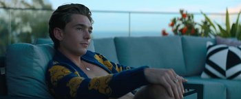 Movie still from “Do Revenge” (2022), directed by Jennifer Kaytin Robinson – A man sitting on top of a couch in a room; Medium shot, Low angle