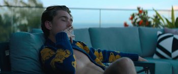 Movie still from “Do Revenge” (2022), directed by Jennifer Kaytin Robinson – A man sitting on a couch eating a sandwich; Medium shot, Low angle