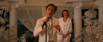 Movie still from “Do Revenge” (2022), directed by Jennifer Kaytin Robinson – A man in a white suit is speaking into a microphone; Medium shot, Over the shoulder angle