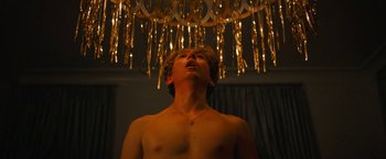 Movie still from “Do Revenge” (2022), directed by Jennifer Kaytin Robinson – A shirtless man standing under a chandelier looking up; Medium shot, Low angle