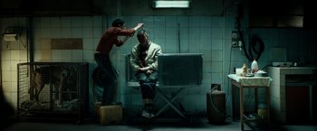 Movie still from “Dogman” (2018), directed by Matteo Garrone – A man sitting on a bench in a room with another man; Wide shot, Over the shoulder angle