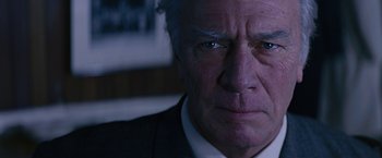 An older man in a suit stares into the camera