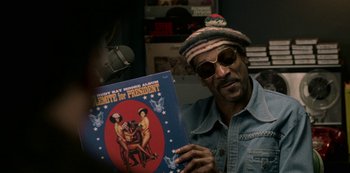 Movie still from “Dolemite Is My Name” (2019), directed by Craig Brewer – A man holding up an album cover in front of a microphone; Medium shot, Over the shoulder angle