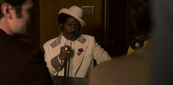 Movie still from “Dolemite Is My Name” (2019), directed by Craig Brewer – A man in a white suit and white hat holding a microphone; Medium shot, Over the shoulder angle