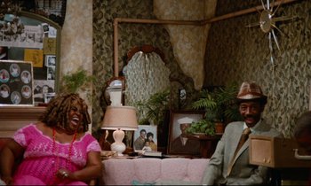 Movie still from “Don't Play Us Cheap” (1972), directed by Melvin Van Peebles – A couple of people sitting on top of a pink couch; Wide shot, High angle