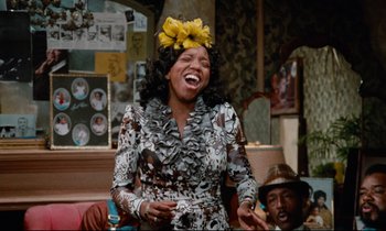 Movie still from “Don't Play Us Cheap” (1972), directed by Melvin Van Peebles – A woman wearing a flower crown and a dress laughing; Medium shot, Low angle