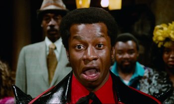 Movie still from “Don't Play Us Cheap” (1972), directed by Melvin Van Peebles – A person wearing a suit and tie; Close Up shot, Low angle