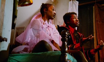 Movie still from “Don't Play Us Cheap” (1972), directed by Melvin Van Peebles – A man and a woman sitting next to each other on top of a bed; Medium shot, Low angle
