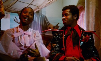 Movie still from “Don't Play Us Cheap” (1972), directed by Melvin Van Peebles – A man and a woman sitting next to each other; Medium shot, Low angle