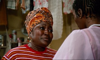 Movie still from “Don't Play Us Cheap” (1972), directed by Melvin Van Peebles – An older woman wearing a head scarf and a red and white striped shirt; Close Up shot, Over the shoulder angle