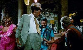 Movie still from “Don't Play Us Cheap” (1972), directed by Melvin Van Peebles – A man in a suit and tie standing next to two other men; Medium shot, Over the shoulder angle