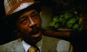 Movie still from “Don't Play Us Cheap” (1972), directed by Melvin Van Peebles – A person wearing a suit and a hat; Close Up shot, Low angle