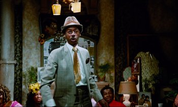 Movie still from “Don't Play Us Cheap” (1972), directed by Melvin Van Peebles – A man in a suit and hat standing in front of a group of people; Medium shot, Low angle