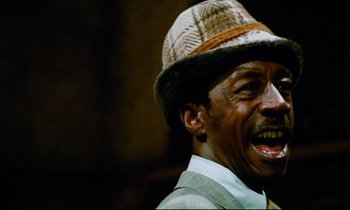 Movie still from “Don't Play Us Cheap” (1972), directed by Melvin Van Peebles – An older man wearing a hat and smiling for the camera; Close Up shot, Low angle