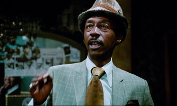 Movie still from “Don't Play Us Cheap” (1972), directed by Melvin Van Peebles – A man wearing a suit and a hat is talking; Close Up shot, Low angle