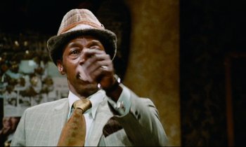 Movie still from “Don't Play Us Cheap” (1972), directed by Melvin Van Peebles – A man in a suit and hat drinking a beer; Close Up shot, Low angle
