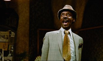 Movie still from “Don't Play Us Cheap” (1972), directed by Melvin Van Peebles – A man wearing a suit and a hat and smiling; Close Up shot, Low angle
