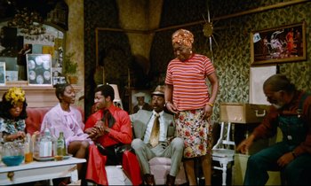 Movie still from “Don't Play Us Cheap” (1972), directed by Melvin Van Peebles – A group of men and women sitting in a living room; Wide shot, High angle