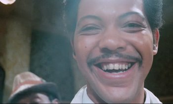 Movie still from “Don't Play Us Cheap” (1972), directed by Melvin Van Peebles – A person with a mustache smiling; Close Up shot, Low angle