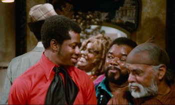 Movie still from “Don't Play Us Cheap” (1972), directed by Melvin Van Peebles – A group of men standing next to each other in a room; Medium shot, Over the shoulder angle