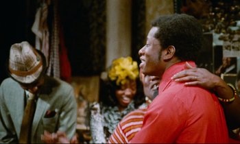 Movie still from “Don't Play Us Cheap” (1972), directed by Melvin Van Peebles – A group of young people laughing together in a room; Medium shot, Over the shoulder angle